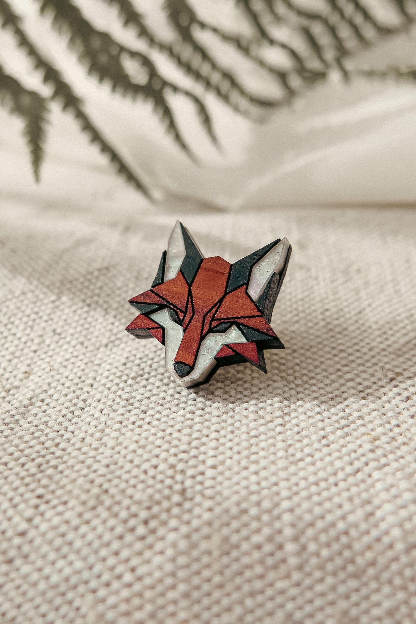 A geometric, handcrafted fox pin made from recycled wood rests on textured beige fabric, with blurred green fern leaves in the background.