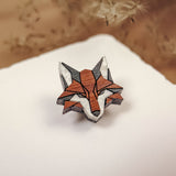 A geometric, origami-style handcrafted fox pin made of recycled wood rests on a white surface, with soft beige plant stems blurred in the background—an eco-conscious accessory with natural charm.