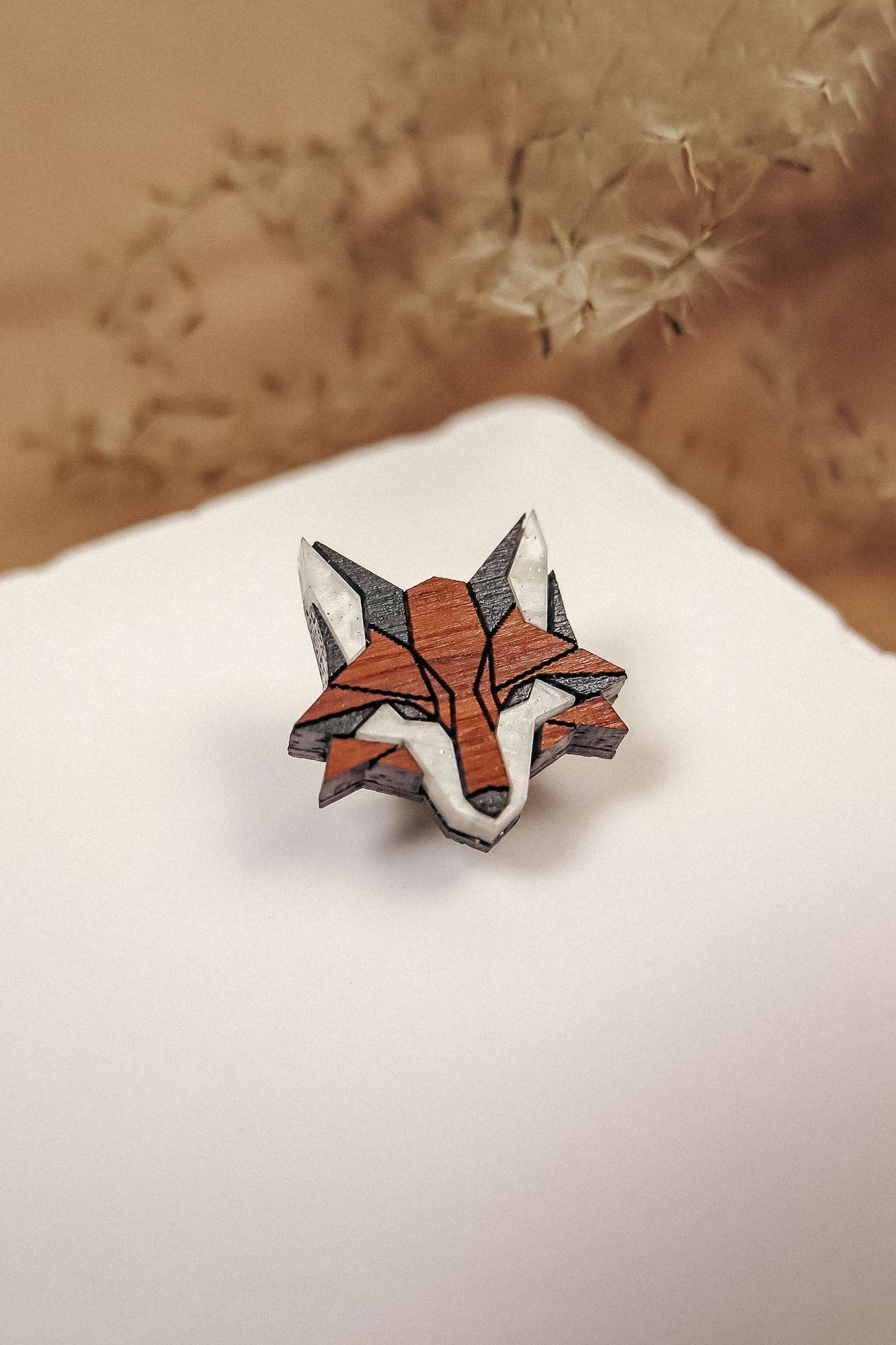 A geometric, origami-style handcrafted fox pin made of recycled wood rests on a white surface, with soft beige plant stems blurred in the background—an eco-conscious accessory with natural charm.