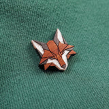 A small, geometric handcrafted fox pin made from recycled wood features orange, brown, and white sections and is pinned to a green textured fabric.