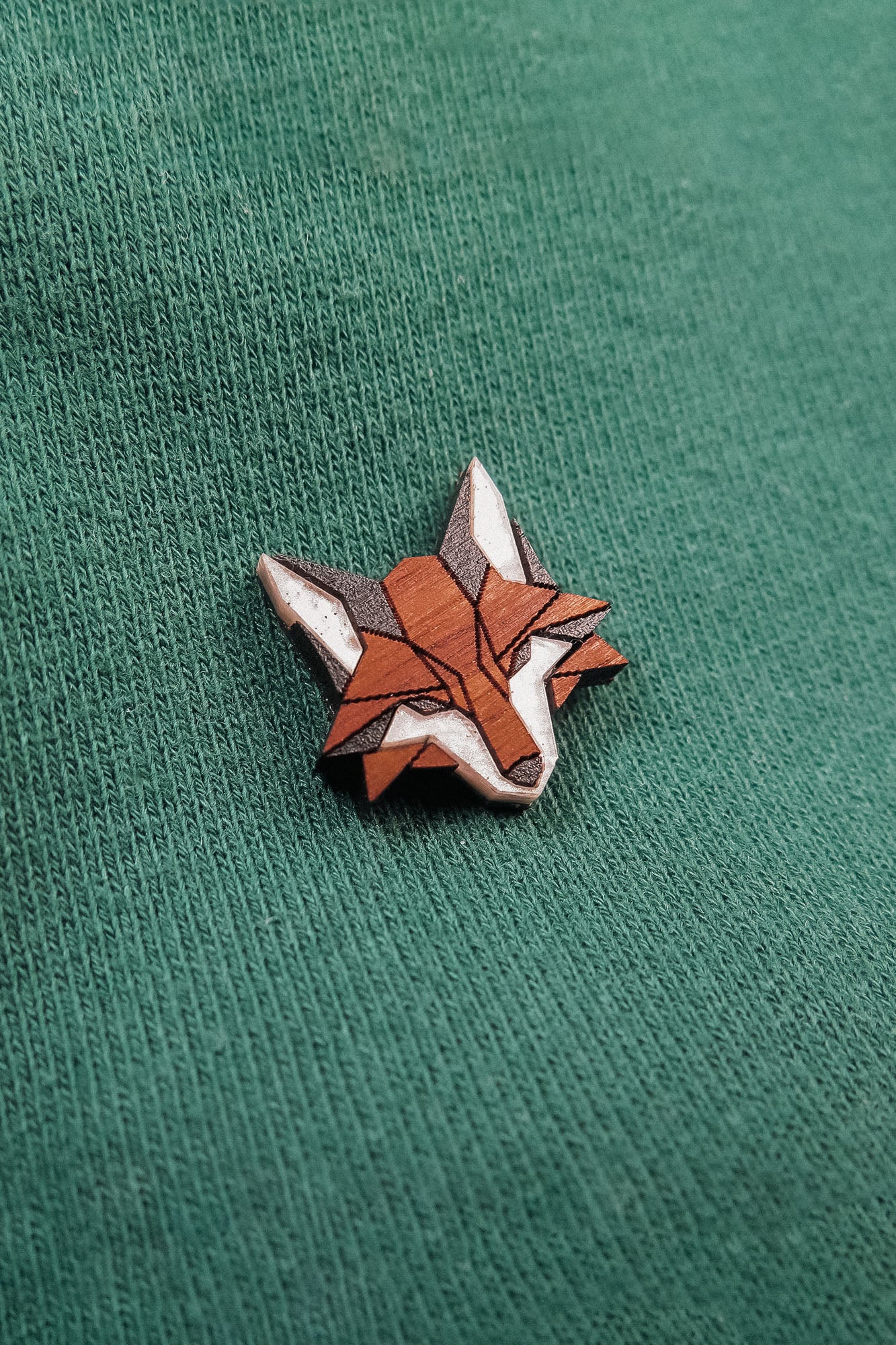 A small, geometric handcrafted fox pin made from recycled wood features orange, brown, and white sections and is pinned to a green textured fabric.