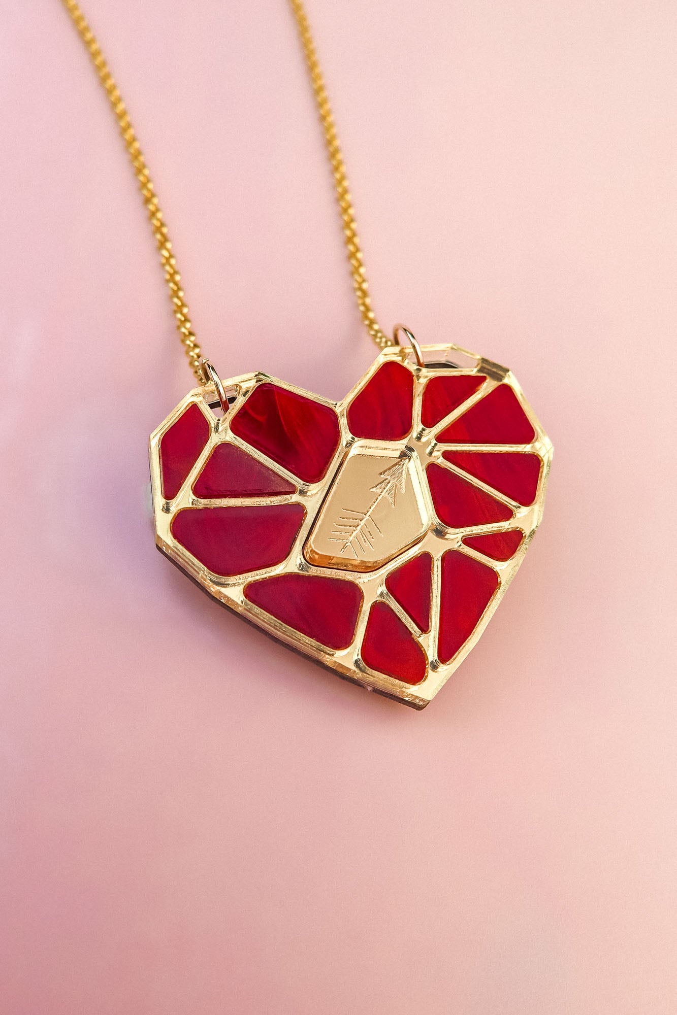 A handcrafted gold chain necklace featuring the Fragment of Love Necklace’s polygon heart pendant, formed from red stained glass outlined in gold, with a central gold plate etched with an arrow, set against a soft pink background.