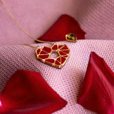 A handcrafted gold Fragment of Love Necklace with a geometric red heart pendant rests on soft pink fabric, surrounded by scattered red rose petals.