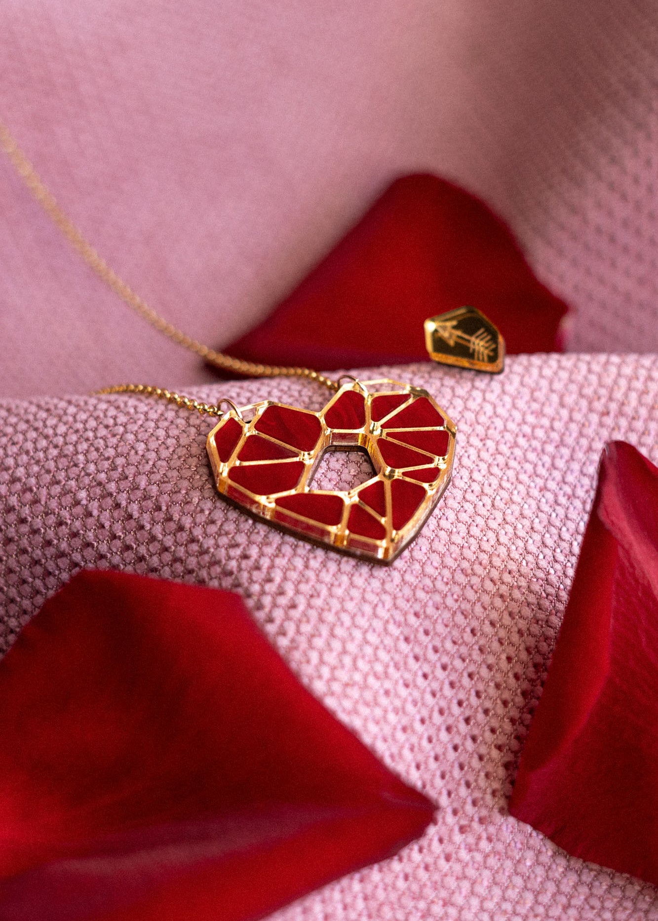 A handcrafted gold Fragment of Love Necklace with a geometric red heart pendant rests on soft pink fabric, surrounded by scattered red rose petals.