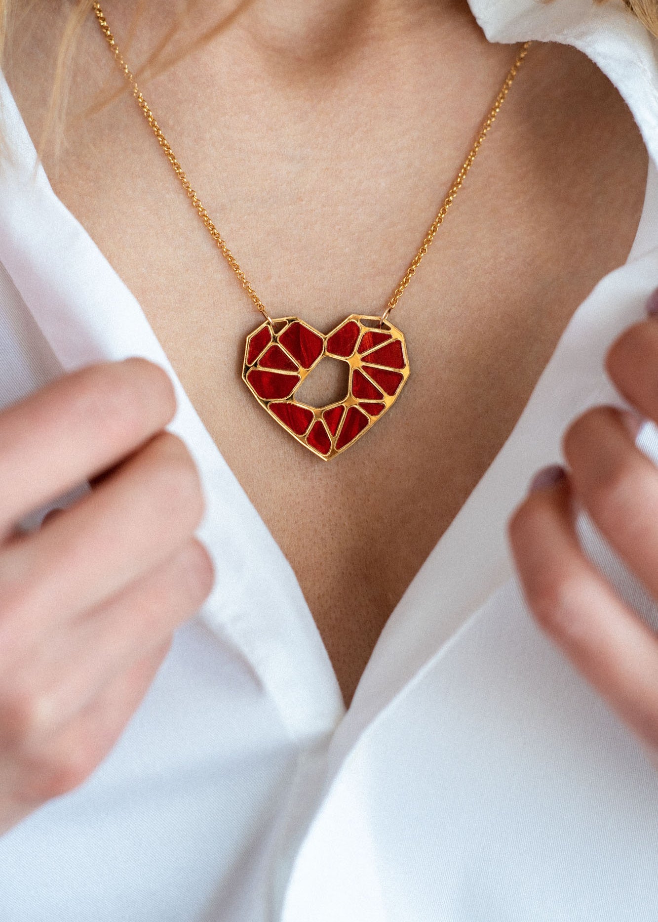 A close-up of a person in a white shirt, holding the collar open to reveal a handcrafted gold necklace with a geometric red heart pendant—an elegant Fragment of Love Necklace displayed around their neck.