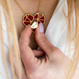 A person with light pink nail polish holds a handcrafted gold and red heart pendant, the Fragment of Love Necklace, against their chest, visible above a white shirt.