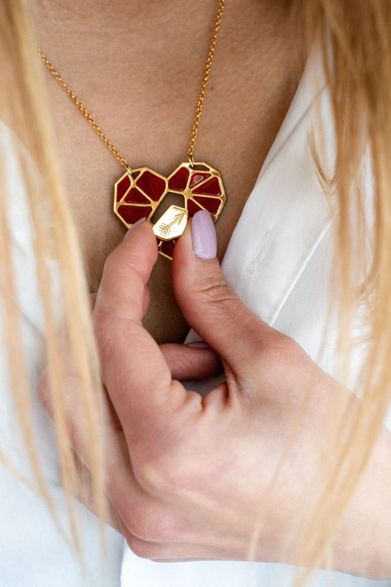 A person with light pink nail polish holds a handcrafted gold and red heart pendant, the Fragment of Love Necklace, against their chest, visible above a white shirt.