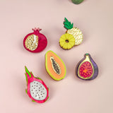 Five colorful fruit-shaped enamel pins—pomegranate, pineapple, papaya, fig, and dragon fruit—are arranged on a light pink surface with green succulent plants at the edges, making a vibrant handcrafted fashion accessory set.