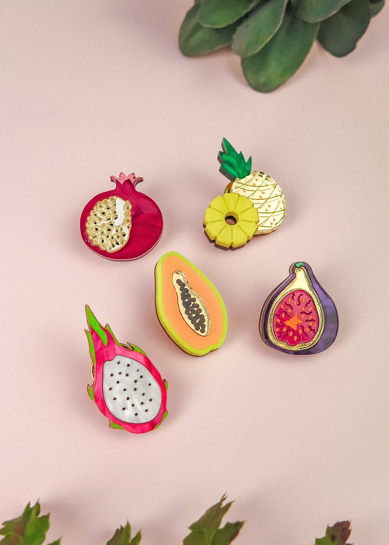Five colorful fruit-shaped enamel pins—pomegranate, pineapple, papaya, fig, and dragon fruit—are arranged on a light pink surface with green succulent plants at the edges, making a vibrant handcrafted fashion accessory set.