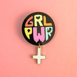 A round black pin with colorful letters spelling GRL PWR and a dangling gold Venus symbol, this handcrafted accessory celebrates meaningful symbolism, set against a vibrant pink background.