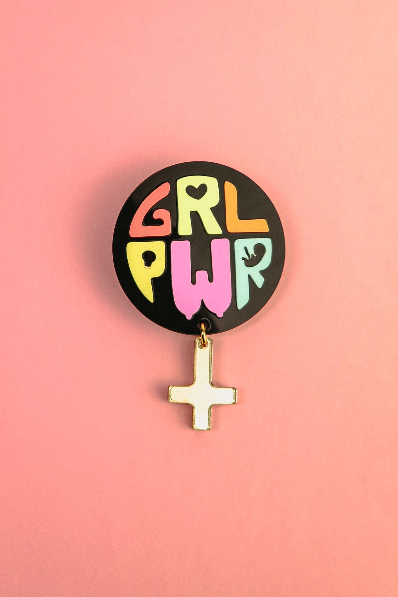 A round black pin with colorful letters spelling GRL PWR and a dangling gold Venus symbol, this handcrafted accessory celebrates meaningful symbolism, set against a vibrant pink background.
