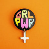 A round Girl Power Brooch with colorful letters spelling GRL PWR and a small white Venus symbol charm hangs below, set against a bright orange background—an expressive handcrafted accessory with meaningful symbolism.