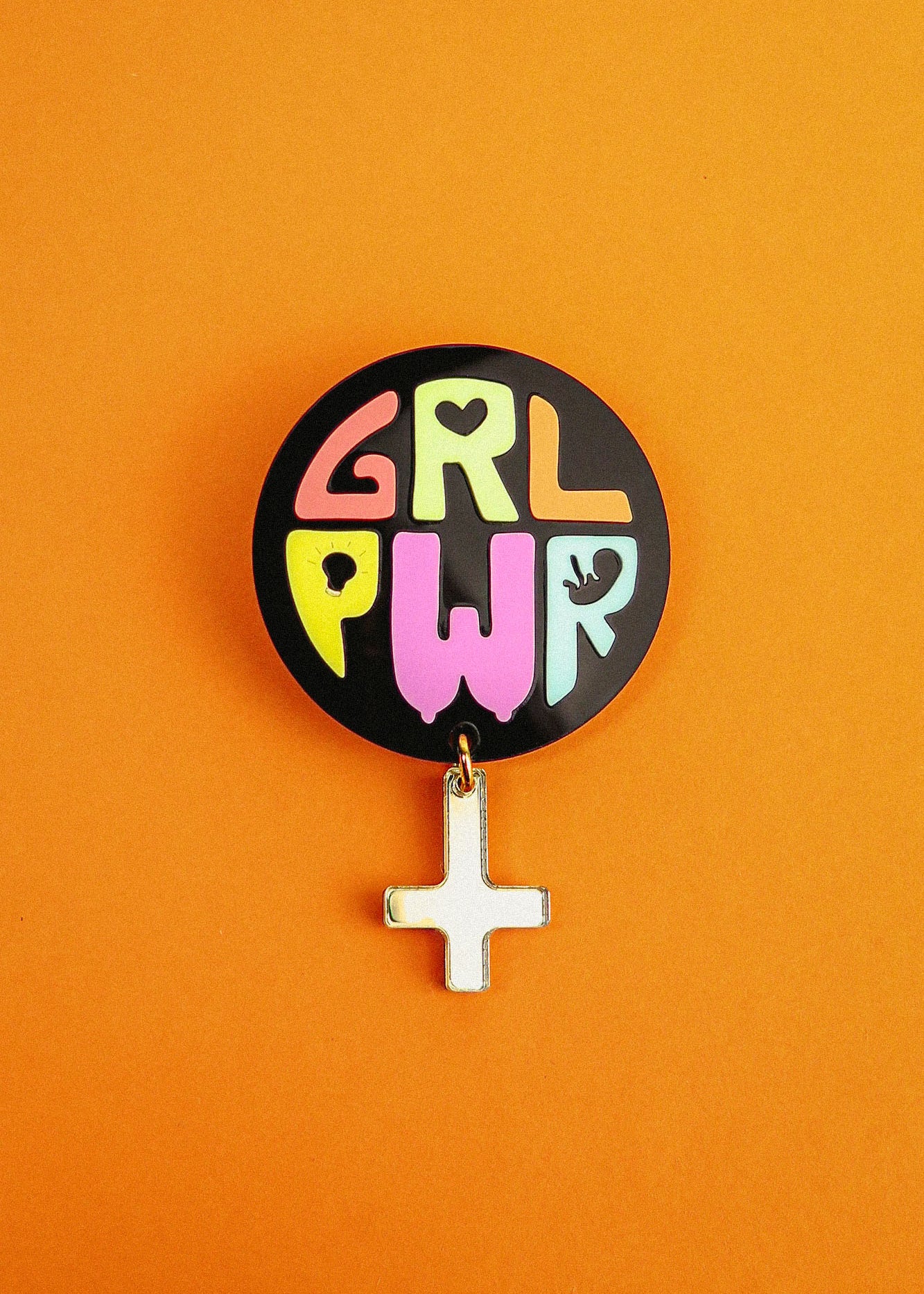 A round Girl Power Brooch with colorful letters spelling GRL PWR and a small white Venus symbol charm hangs below, set against a bright orange background—an expressive handcrafted accessory with meaningful symbolism.