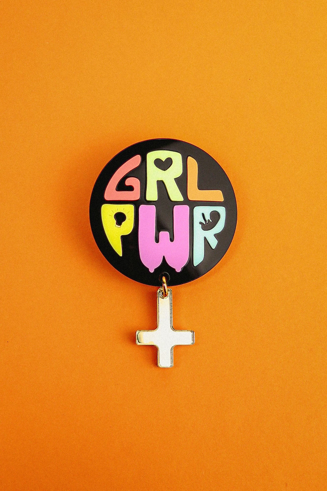 A round Girl Power Brooch with colorful letters spelling GRL PWR and a small white Venus symbol charm hangs below, set against a bright orange background—an expressive handcrafted accessory with meaningful symbolism.