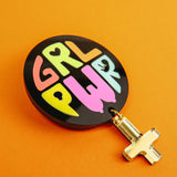 A round black keychain with GRL PWR written in colorful, playful letters, attached to a gold gender symbol charm, lies on a bright orange surface—a handcrafted accessory full of meaningful symbolism.