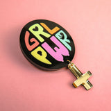 A round black Girl Power Brooch with GRL PWR in colorful letters and a gold female gender symbol charm hanging below, placed on a pink background—a handcrafted accessory celebrating meaningful symbolism.