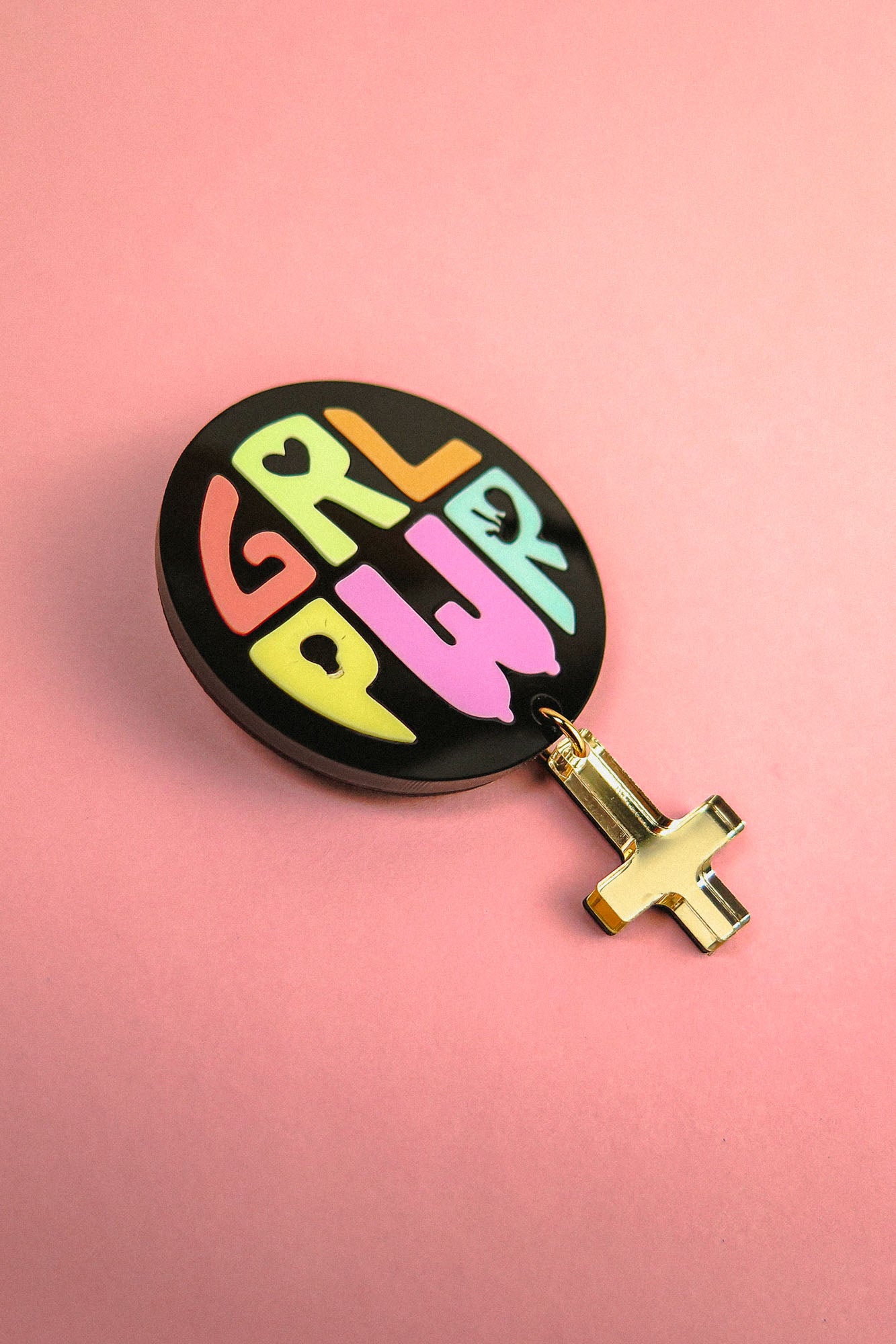 A round black Girl Power Brooch with GRL PWR in colorful letters and a gold female gender symbol charm hanging below, placed on a pink background—a handcrafted accessory celebrating meaningful symbolism.