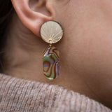 Close-up of a person’s ear wearing a handcrafted gold earring with a sunburst design, featuring a dangling abstract, multicolored abalone piece. The person is wearing a taupe knit sweater.