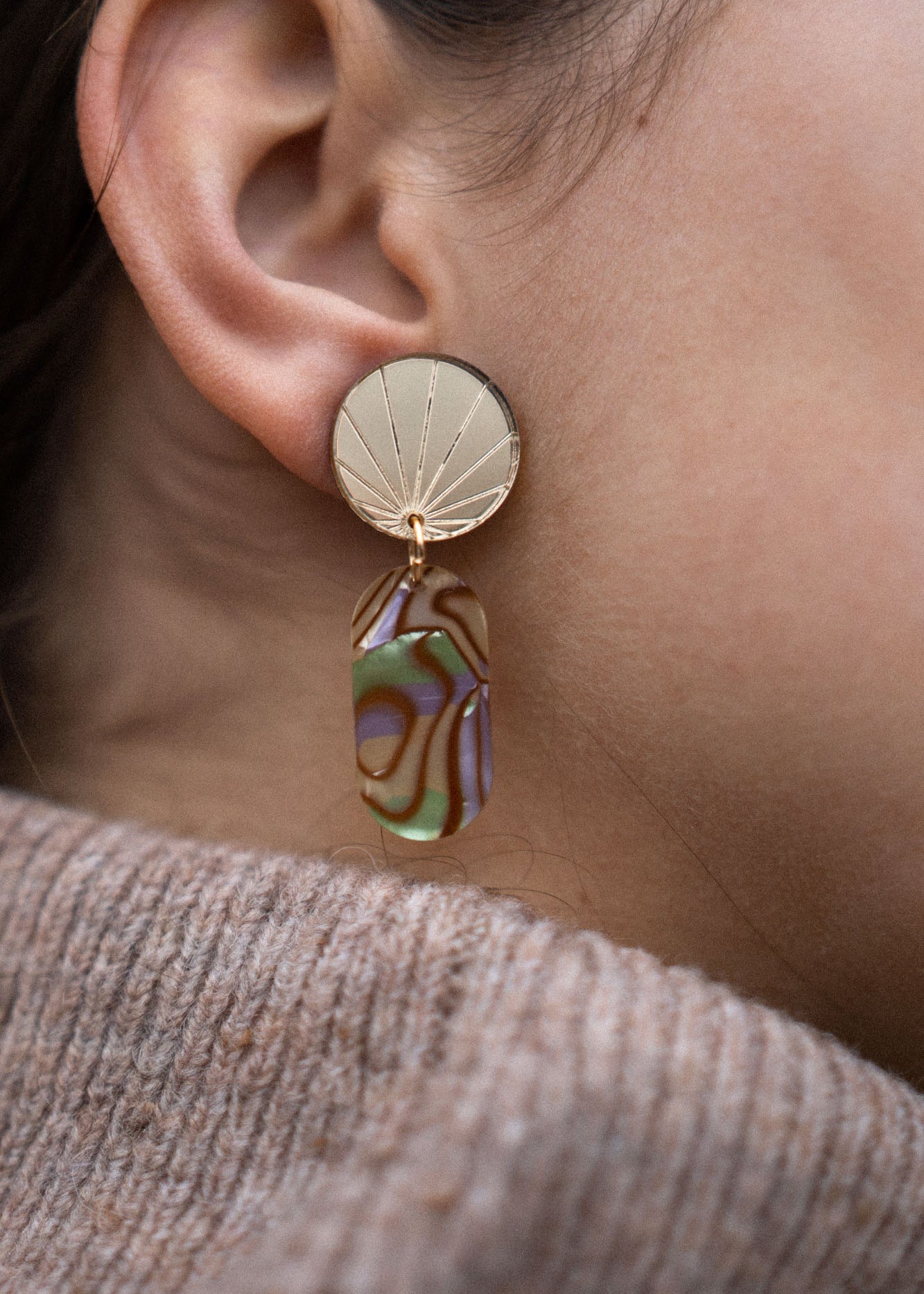 Close-up of a person’s ear wearing a handcrafted gold earring with a sunburst design, featuring a dangling abstract, multicolored abalone piece. The person is wearing a taupe knit sweater.