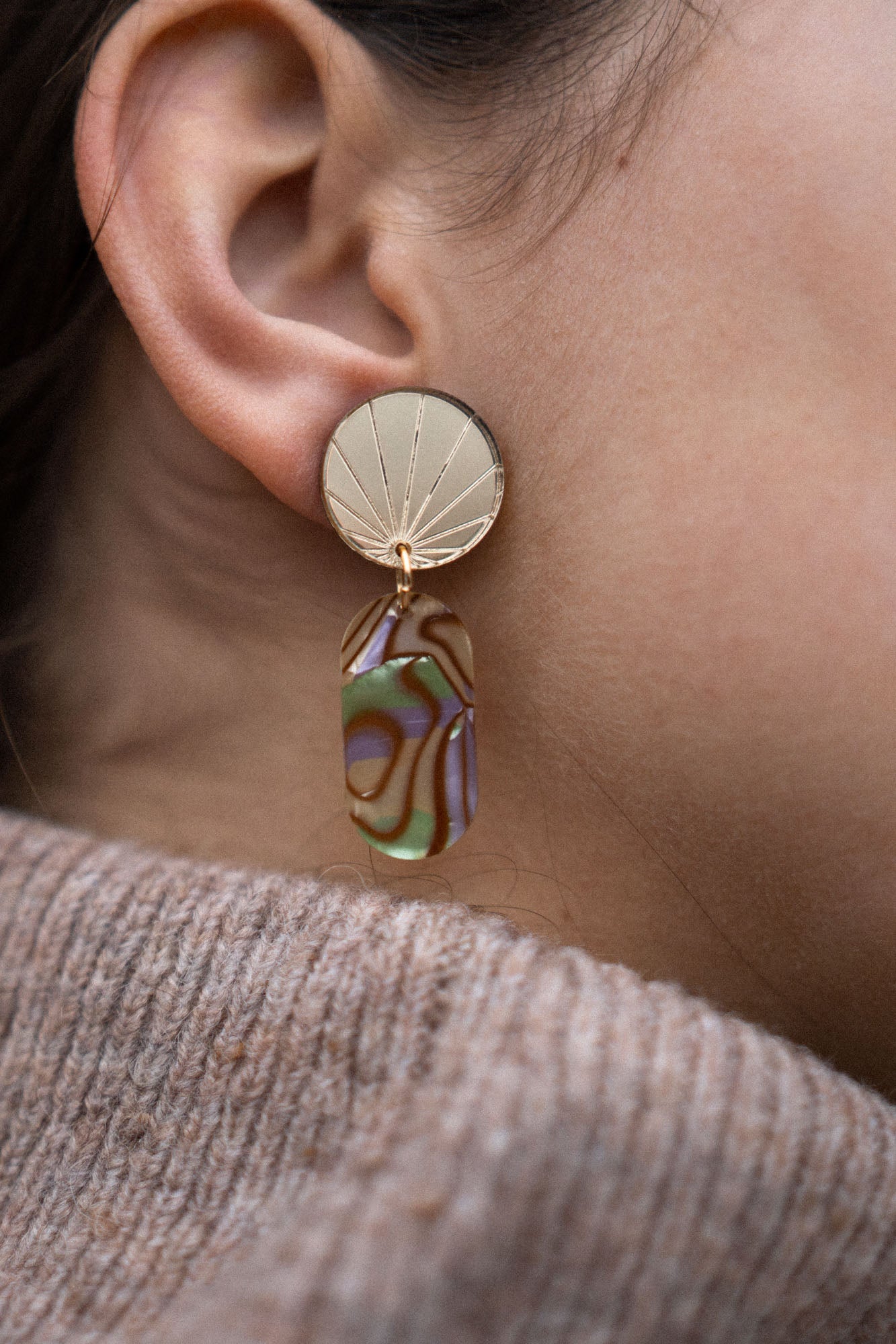 Close-up of a person’s ear wearing a handcrafted gold earring with a sunburst design, featuring a dangling abstract, multicolored abalone piece. The person is wearing a taupe knit sweater.