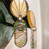 Close-up of handcrafted gold earrings with engraved fan-patterned tops and elongated oval dangles made of translucent abalone, showcasing wavy green, brown, and cream stripes.