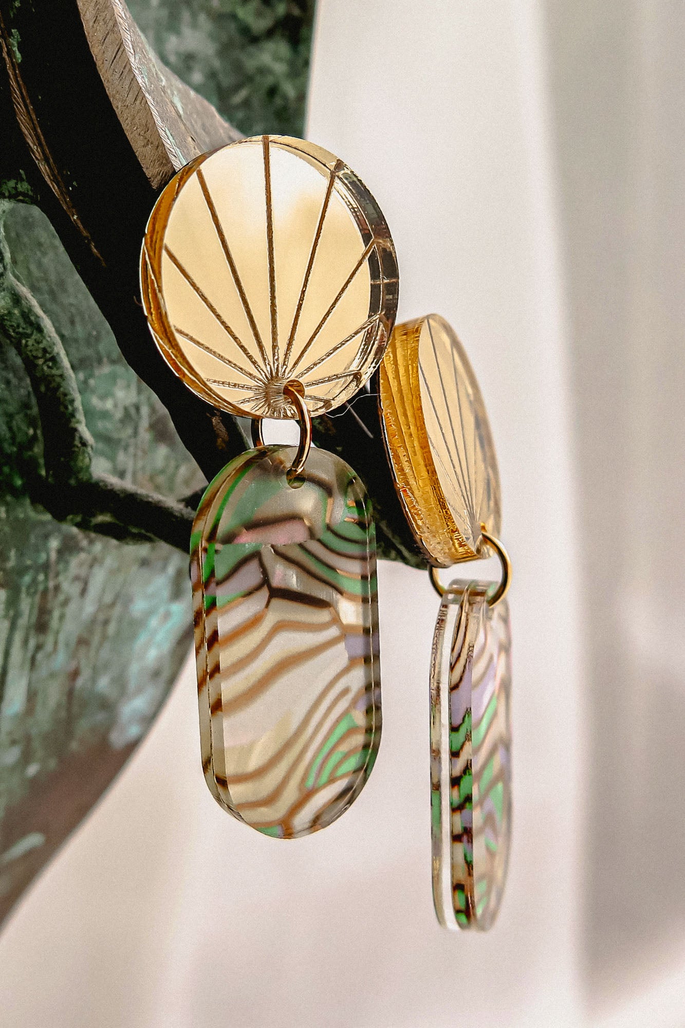 Close-up of handcrafted gold earrings with engraved fan-patterned tops and elongated oval dangles made of translucent abalone, showcasing wavy green, brown, and cream stripes.