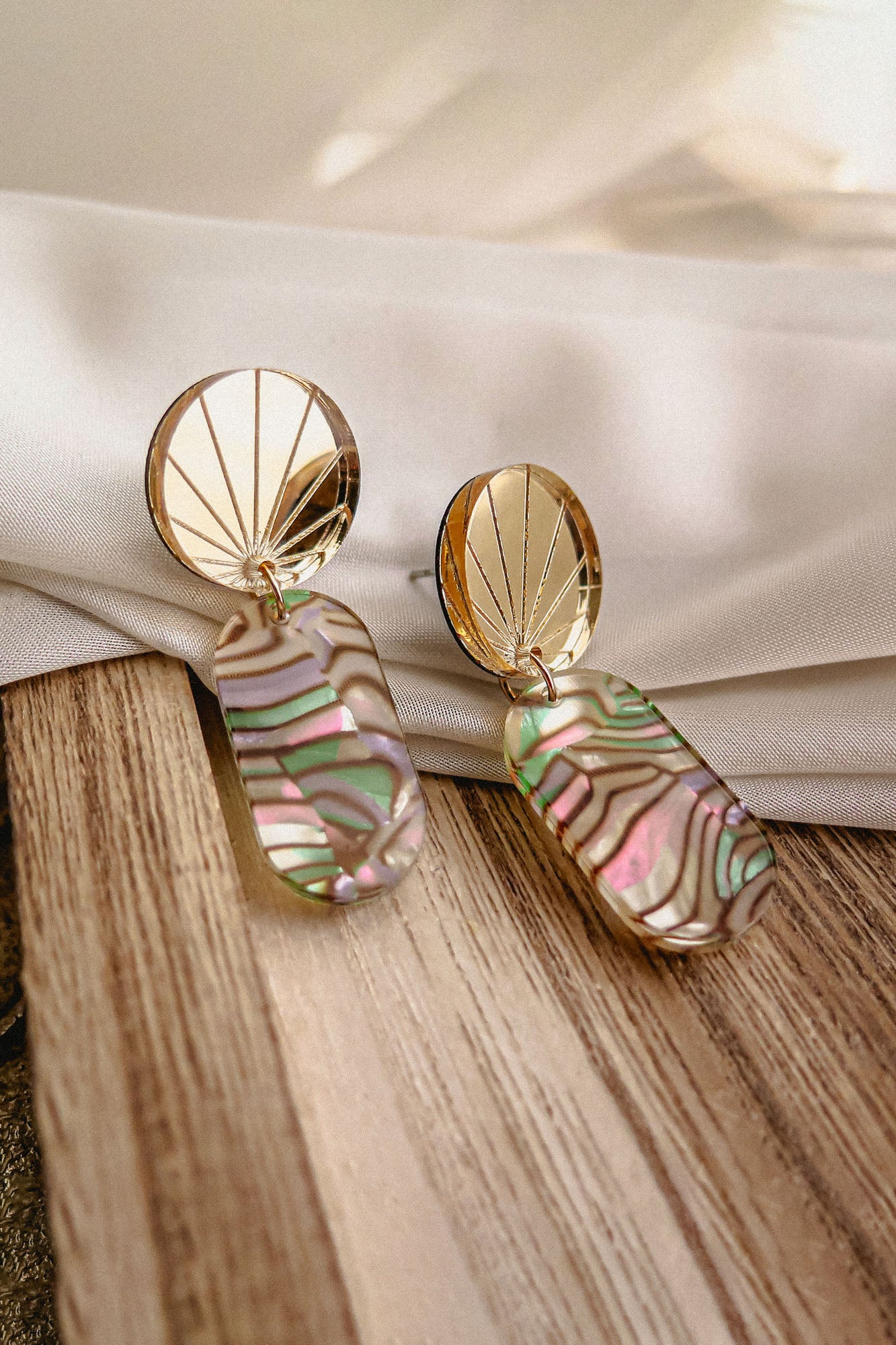 A pair of handcrafted gold earrings with fan-shaped tops and oval, iridescent abalone shell pendants rests on a light wood surface, with a white fabric backdrop in soft natural light.
