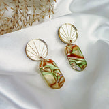 Two handcrafted gold earrings rest on white satin fabric. Each features a sunburst design and a dangling rectangular pendant with marbled green, brown, and cream hues, reminiscent of abalone. Delicate dried foliage lies nearby.