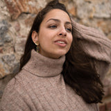 A woman in a beige turtleneck sweater stands against a textured stone wall, looking slightly to the side with a relaxed expression, one hand in her hair, and handcrafted gold earrings catching the light.