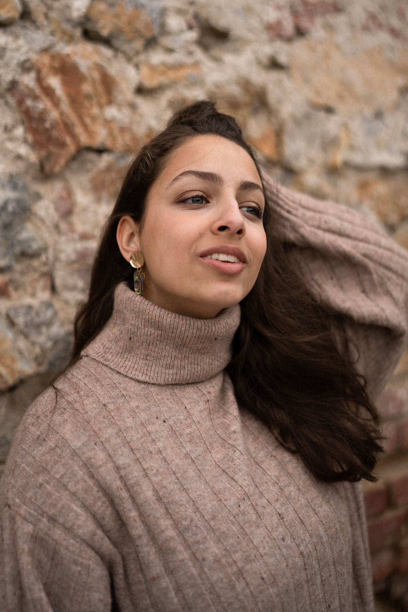 A woman in a beige turtleneck sweater stands against a textured stone wall, looking slightly to the side with a relaxed expression, one hand in her hair, and handcrafted gold earrings catching the light.