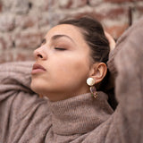 A woman wearing a beige turtleneck sweater and handcrafted gold earrings poses with her eyes closed, head tilted back, and hands resting behind her head in front of a brick wall.