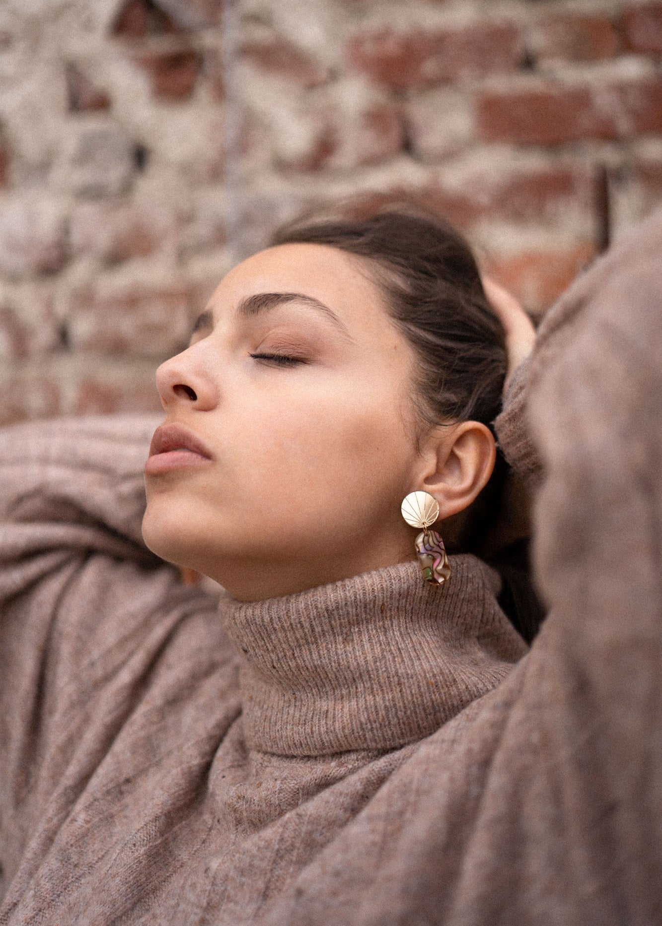 A woman wearing a beige turtleneck sweater and handcrafted gold earrings poses with her eyes closed, head tilted back, and hands resting behind her head in front of a brick wall.