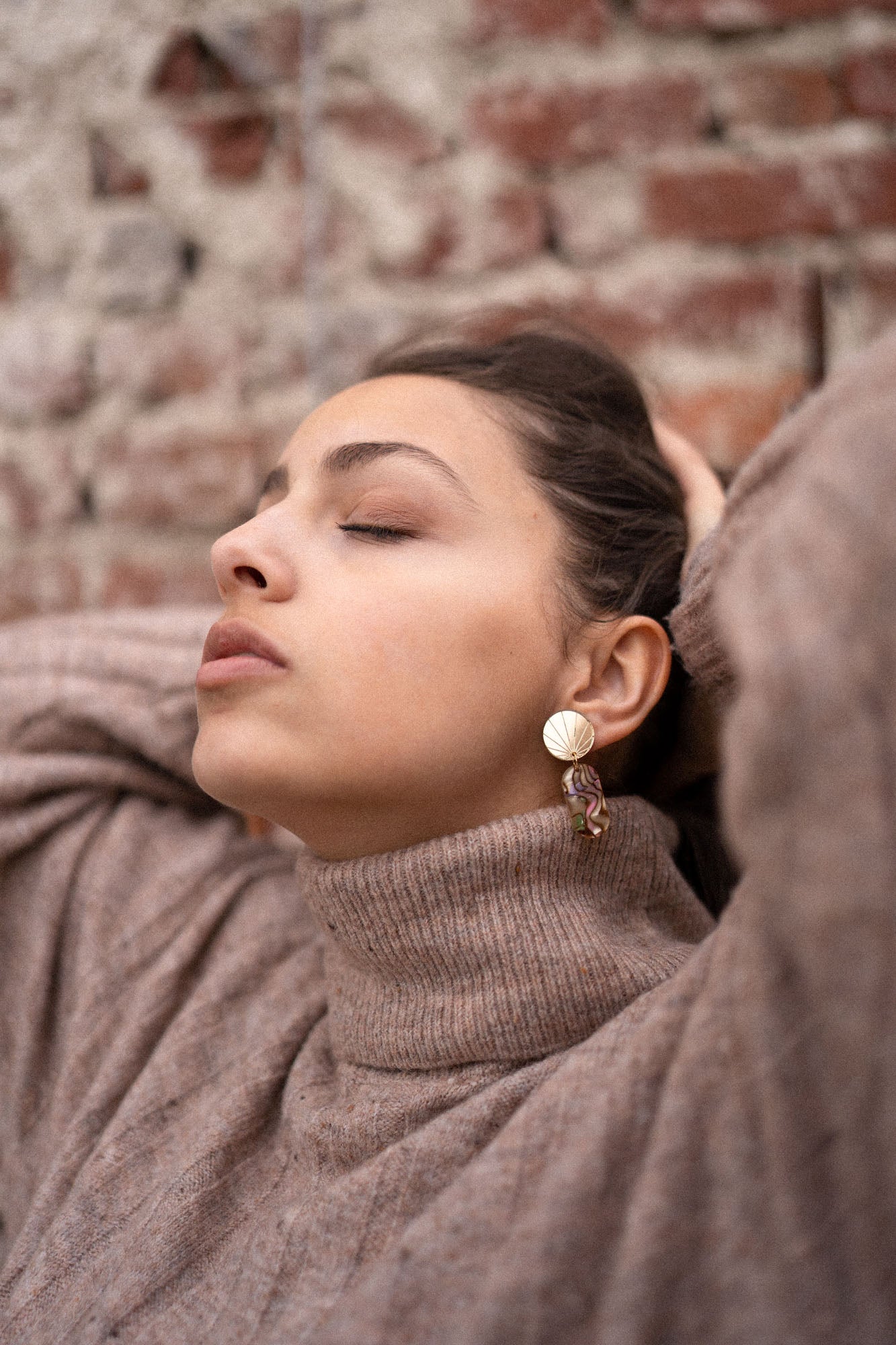 A woman wearing a beige turtleneck sweater and handcrafted gold earrings poses with her eyes closed, head tilted back, and hands resting behind her head in front of a brick wall.