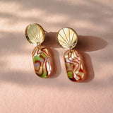 A pair of handcrafted gold earrings featuring round, fan-patterned tops and rectangular, abalone-inspired marbled pendants in green, pink, and cream tones, displayed on a soft pink surface with gentle shadows.