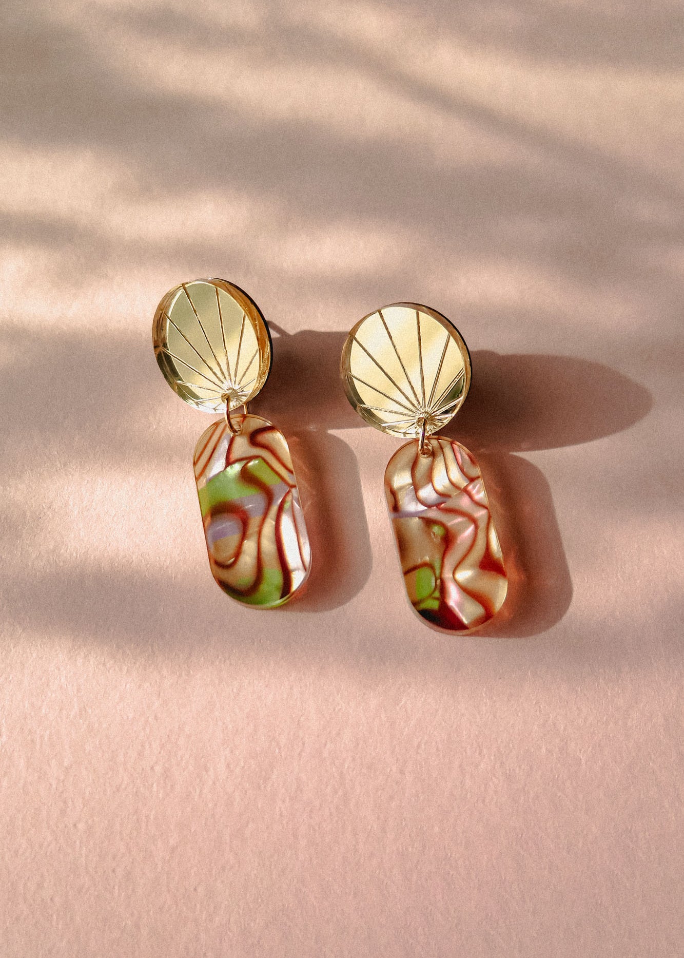 A pair of handcrafted gold earrings featuring round, fan-patterned tops and rectangular, abalone-inspired marbled pendants in green, pink, and cream tones, displayed on a soft pink surface with gentle shadows.