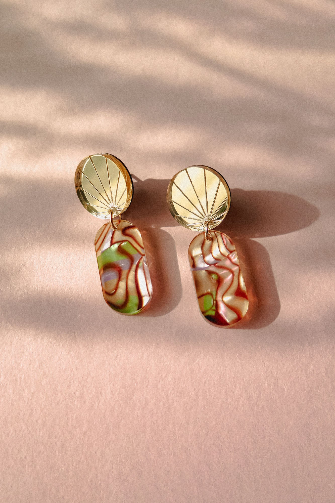 A pair of handcrafted gold earrings featuring round, fan-patterned tops and rectangular, abalone-inspired marbled pendants in green, pink, and cream tones, displayed on a soft pink surface with gentle shadows.