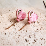 A pair of pink Heart Balloon Studs with gold accents and dangling gold chains, displayed on a textured, light-colored surface—perfect for adding vibrant style to any look.