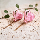 A pair of pink Heart Balloon Studs with gold accents and dangling chains, displayed on a textured cream surface with a green leafy stem in the background—perfect for adding vibrant style to any look.
