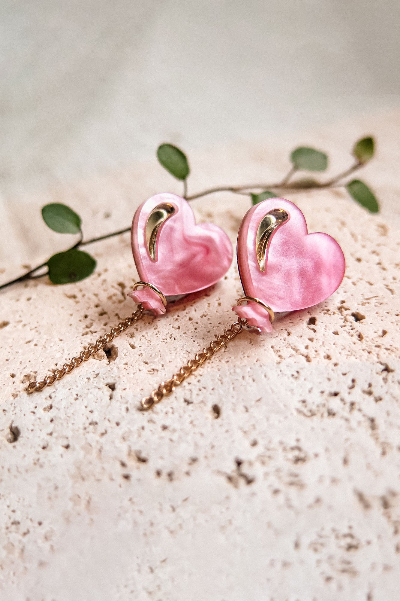 A pair of pink Heart Balloon Studs with gold accents and dangling chains, displayed on a textured cream surface with a green leafy stem in the background—perfect for adding vibrant style to any look.