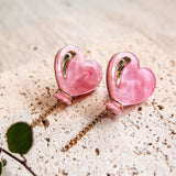 A pair of pink Heart Balloon Studs with gold accents and dangling gold chains, displayed on a textured, light-colored stone surface. A small green plant is visible in the lower left corner, completing this vibrant style.