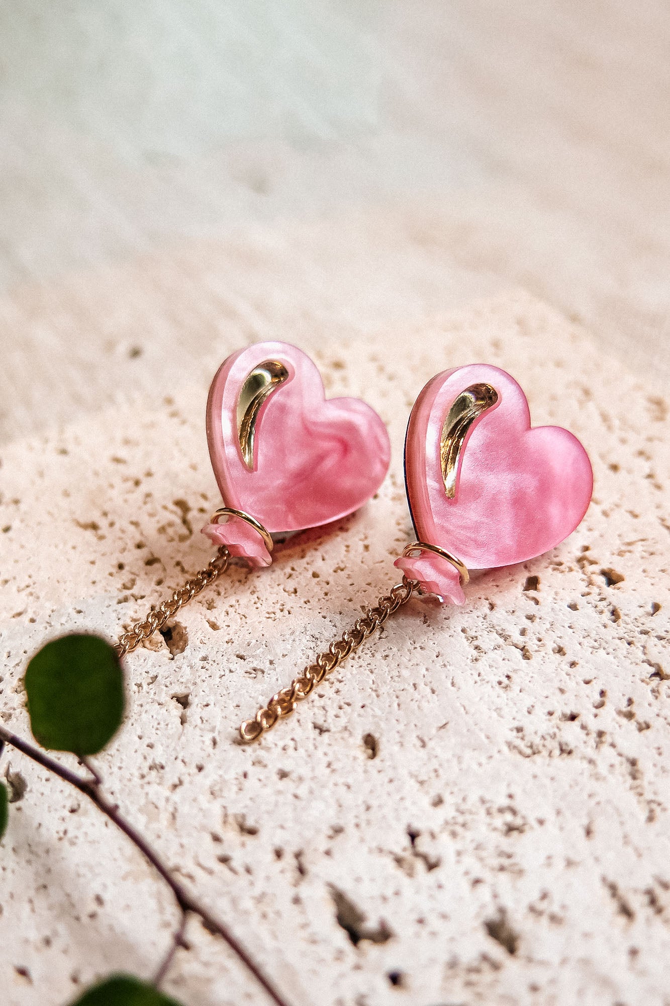 A pair of pink Heart Balloon Studs with gold accents and dangling gold chains, displayed on a textured, light-colored stone surface. A small green plant is visible in the lower left corner, completing this vibrant style.