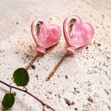 A pair of pink Heart Balloon Studs, heart-shaped earrings with gold accents and dangling gold chains, displayed on a textured beige surface. A green leafy stem lies nearby, adding to their vibrant style.