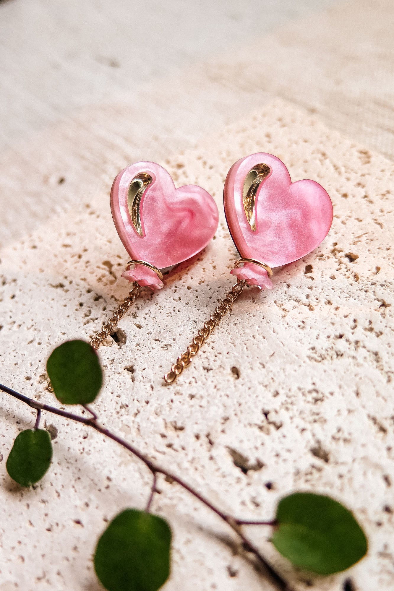 A pair of pink Heart Balloon Studs, heart-shaped earrings with gold accents and dangling gold chains, displayed on a textured beige surface. A green leafy stem lies nearby, adding to their vibrant style.