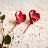 A pair of Heart Balloon Studs with a playful, vibrant style—red heart-shaped earrings featuring gold detailing and delicate chains, displayed on a textured beige surface beside a small green leafy plant.