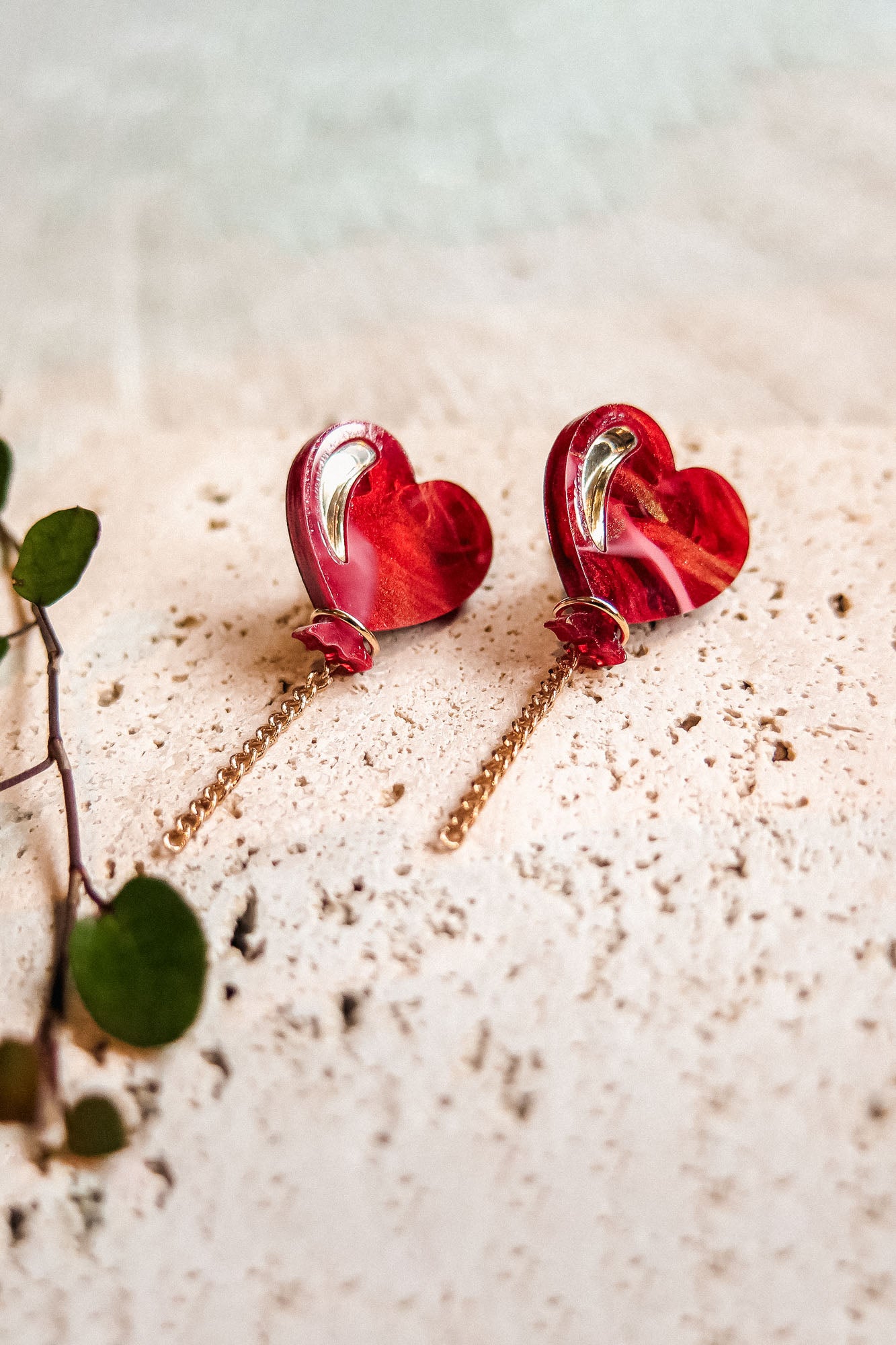 A pair of Heart Balloon Studs with a playful, vibrant style—red heart-shaped earrings featuring gold detailing and delicate chains, displayed on a textured beige surface beside a small green leafy plant.