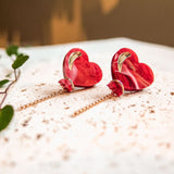 A pair of Heart Balloon Studs—red, heart-shaped earrings with gold accents and delicate chain drops—are displayed on a speckled white surface, exuding a playful, vibrant style as a green vine creeps into the blurred background.