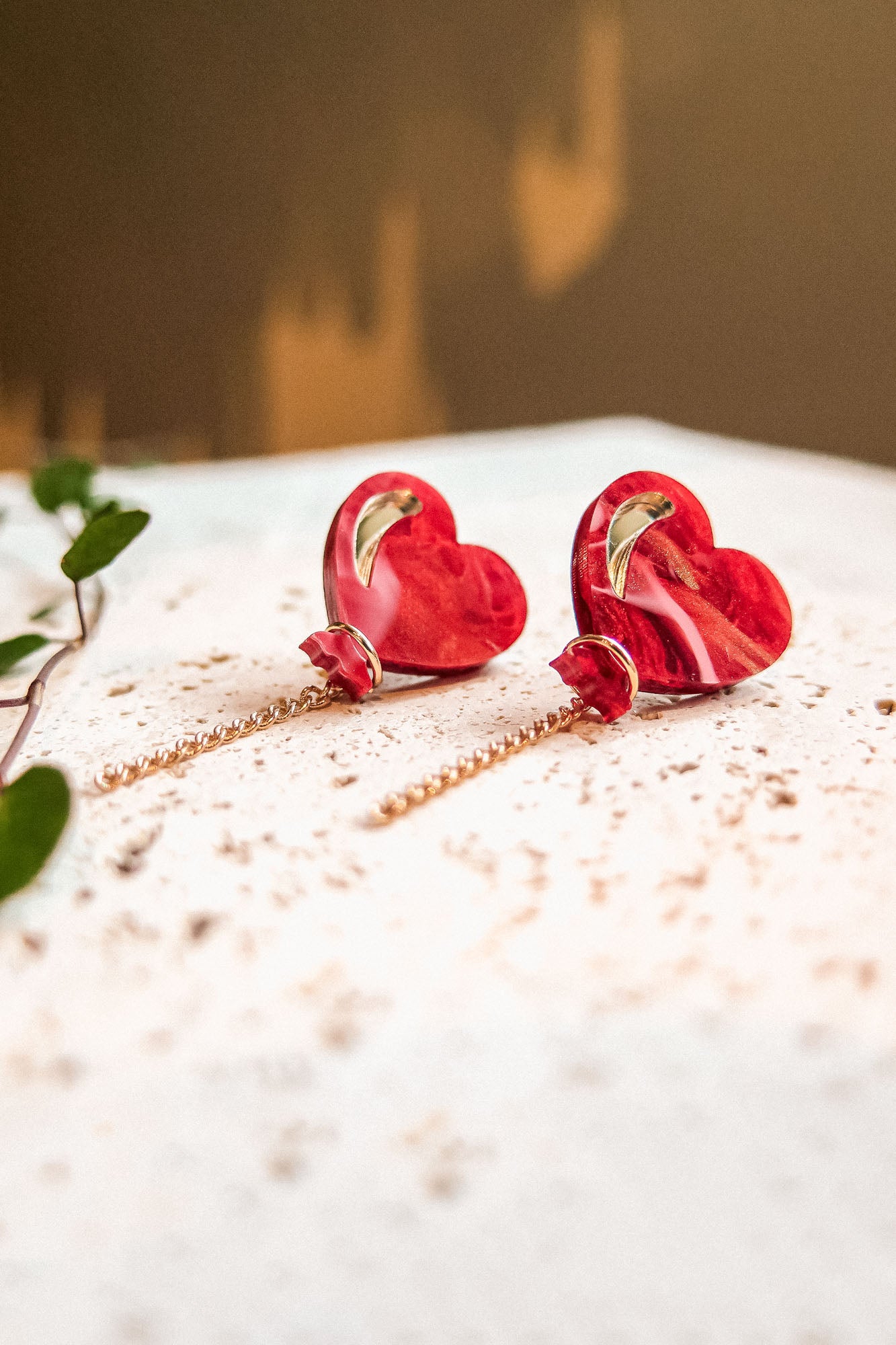 A pair of Heart Balloon Studs—red, heart-shaped earrings with gold accents and delicate chain drops—are displayed on a speckled white surface, exuding a playful, vibrant style as a green vine creeps into the blurred background.