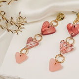 A pair of Heart Chain Earrings with gold links and pink heart-shaped charms, displayed on a white textured surface with delicate dried flowers and draped ivory fabric in the background, adds a touch of allergy-free comfort to your look.