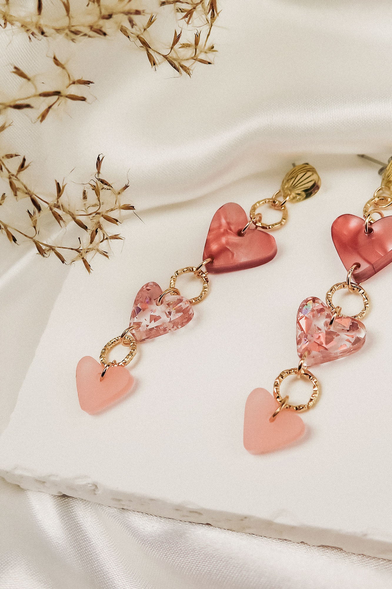 A pair of Heart Chain Earrings with gold links and pink heart-shaped charms, displayed on a white textured surface with delicate dried flowers and draped ivory fabric in the background, adds a touch of allergy-free comfort to your look.