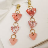 A pair of Heart Chain Earrings featuring three heart-shaped charms in pink tones, linked with gold rings for allergy-free comfort, displayed on a white textured surface with a satin fabric background.